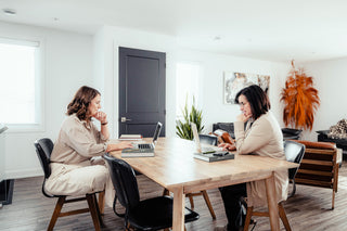 Two women sitting at a wooden table in a modern office setting, using laptops.