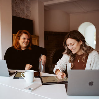 Two women sitting at a table with laptops and a tablet, smiling.